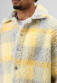 Gele en grijze geruite fleecejas met knoopsluiting, gestructureerde stof en een kraag. Zacht materiaal met zichtbare stiksels.