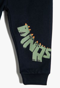 Granatowe dresy z zielonym grafiką dinozaura z pomarańczowymi kolcami oraz napisem "DINOS" w zabawnym foncie. Gładka faktura materiału, ściągacz na dole.