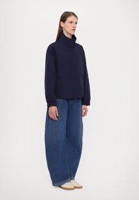 Navy blue cropped jacket with a high collar, elastic cuffs, and two front pockets, paired with wide-leg denim jeans and beige shoes.