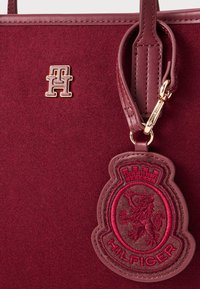 Burgunderfarbene Tragetasche aus strukturiertem Stoff mit einem goldenen Logo und einem roten gestickten Etikett mit einem Löwen- und Kronenmotiv.