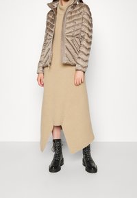 Person in einem beige-gerippten Strickkleid mit asymmetrischem Saum, einer hellbraunen, gesteppten Jacke und schwarzen Schnürkampfstiefeln vor einem weißen Hintergrund.