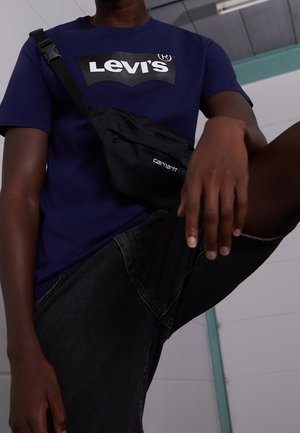 Person iført en Marineblå Levi's skjorte, sorte denim shorts og en sort Carhartt crossbody taske, som poser med det ene ben hævet indendørs.