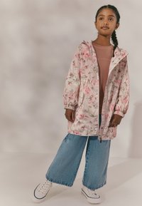 Jeune fille aux cheveux tressés portant une veste rose à fleurs, un haut mauve, un jean bleu à jambes larges et des baskets blanches, debout devant un fond uni.