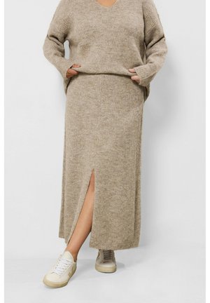 Personne portant un pull en maille beige et une longue jupe assortie avec une fente à l’avant, associée à des baskets blanc cassé, debout sur un fond blanc.