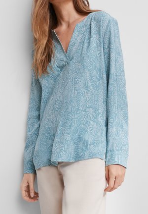 Femme portant un chemisier à manches longues à motifs bleu clair avec un col en V, associé à un pantalon beige clair sur un fond uni.