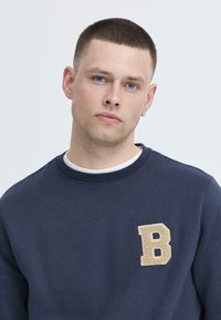 Marineblaues Sweatshirt mit Rundhalsausschnitt, ausgestattet mit einem strukturierten beigen "B"-Aufnäher auf der linken Brust. Glattes Material mit normaler Passform.