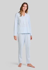 Mujer de pie descalza, vistiendo un conjunto de pijama de manga larga azul claro con cuello abotonado y pantalones holgados, frente a un fondo gris claro.