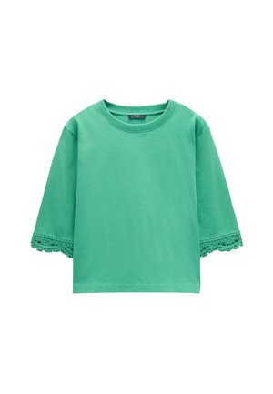 Zielona koszulka z krótkim rękawem, okrągłym dekoltem i koronkowym wykończeniem na krawędziach rękawów, wyłożona na płasko na białym tle.