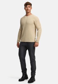 Pull long à manches longues beige en maille côtelée avec un col rond, associé à un jean gris foncé et des bottes noires. Tissu texturé avec une coupe ajustée.
