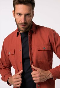 Rotes Baumwollhemd mit Druckknöpfen, zwei Fronttaschen und klassischem Kragen, über einem schwarzen T-Shirt getragen. Besitzt eine glatte Textur.