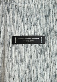 Tela a rayas gris y blanca con un acabado texturizado; cuenta con una etiqueta de cuero negro con texto blanco que detalla la marca y la agencia de diseño.