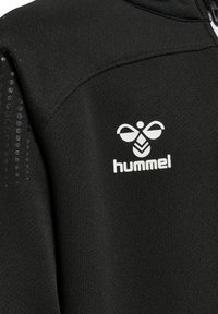 Svart idrottsjacka med texturerat tyg, dragkedja och en framträdande vit "hummel"-logotyp på bröstet. Tilläggsdetaljer på axeln.
