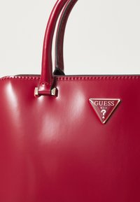 Borsa a mano rossa lucida con texture liscia, dotata di due manici e un accento triangolare in argento sul fronte. Borchie argentate sulle cinghie.