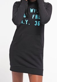Kobieta w czarnej sukience z długim rękawem typu hoodie, z przednią kieszenią, z jasnoniebieskim napisem częściowo brzmiącym "WILD FREE N.Y. 36".