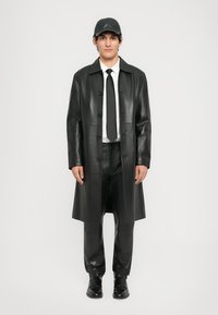 Black leather trench coat with a straight cut, button closure, and long sleeves, worn over black pants and a white shirt, finished with a tie and cap.