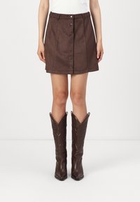 Kvinna iklädd en brun mockaskjorta med knappar framtill, i kombination med höga, mörkbruna cowboystövlar, stående mot en plain vit bakgrund.