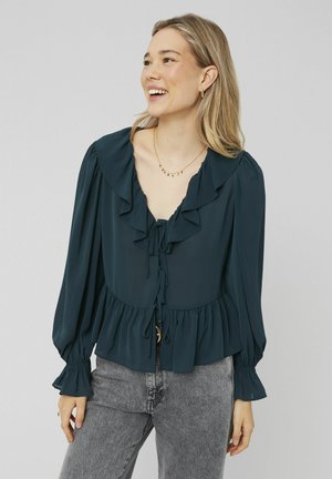 Blouse turquoise avec col et manches volantés, avec un devant noué et une taille peplum. Fabriquée en tissu léger, associée à un jean gris.