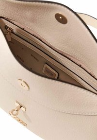 Sac à main en cuir synthétique crème avec une forme structurée, comprenant un accent métallique doré en forme de clé, une poche zippée et une doublure intérieure de marque.