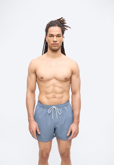 Μυώδης άνδρας με dreadlocks στέκεται προς τα εμπρός φορώντας γκρι μαγιό με κορδόνι, με φόντο απλό.