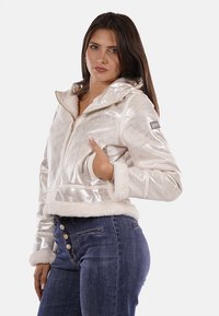 Giacca corta argentata e lucida con bordo in pelliccia sintetica, chiusura con zip e strisce decorative. Abbinata a jeans blu scuro con bottoni ornamentali.