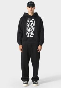 Sudadera negra con un gráfico floral blanco, combinada con pantalones negros holgados y zapatillas negras con suela blanca. Accesorios con pulseras.