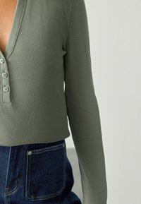 Green ribbed long-sleeve top featuring a deep V-neck and four-button closure, paired with dark blue denim high-waisted pants.