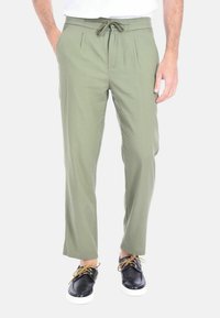 Pantaloni leggeri color verde oliva con vita regolabile tramite cordoncino, con plissettature frontali e gambe dritte. Abbinati a scarpe nere con stringhe.