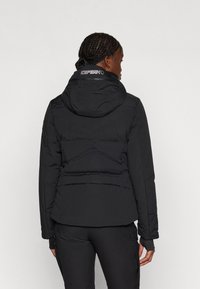 Melna polsterēta jaka ar kapuci, aprīkota ar regulējamu apkakli, izšūtu dizainu un sānu kabatām. Gluda tekstūra ar matētu apdari.