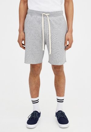 Mężczyzna ubrany w jasnoszare szorty ze sznurkiem, białe skarpetki z czarnymi paskami i granatowe trampki, stojący na tle jednolitego białego tła.