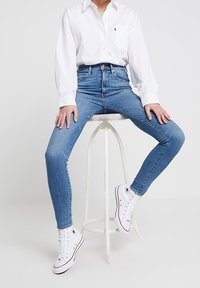 Jeans de mezclilla azul de tiro alto combinados con una camisa blanca de botones y zapatillas blancas, sentada en un taburete blanco.