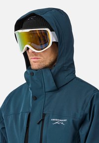 Teal sneeuwjack met een ritssluiting zak, hoge kraag en verstelbare capuchon. Draagt witte monturen met gele glazen en een zwarte beanie.