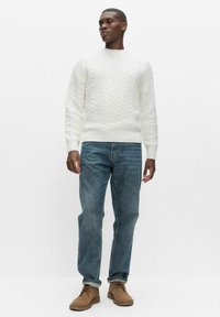 Mies seisoo valkoisessa kaapelineulospuserossa, sinisissä resorikäänteillä varustetuissa farkuissa ja ruskeissa mokkanahkaisissa nauhallisissa saappaissa yksinkertaisen taustan edessä.