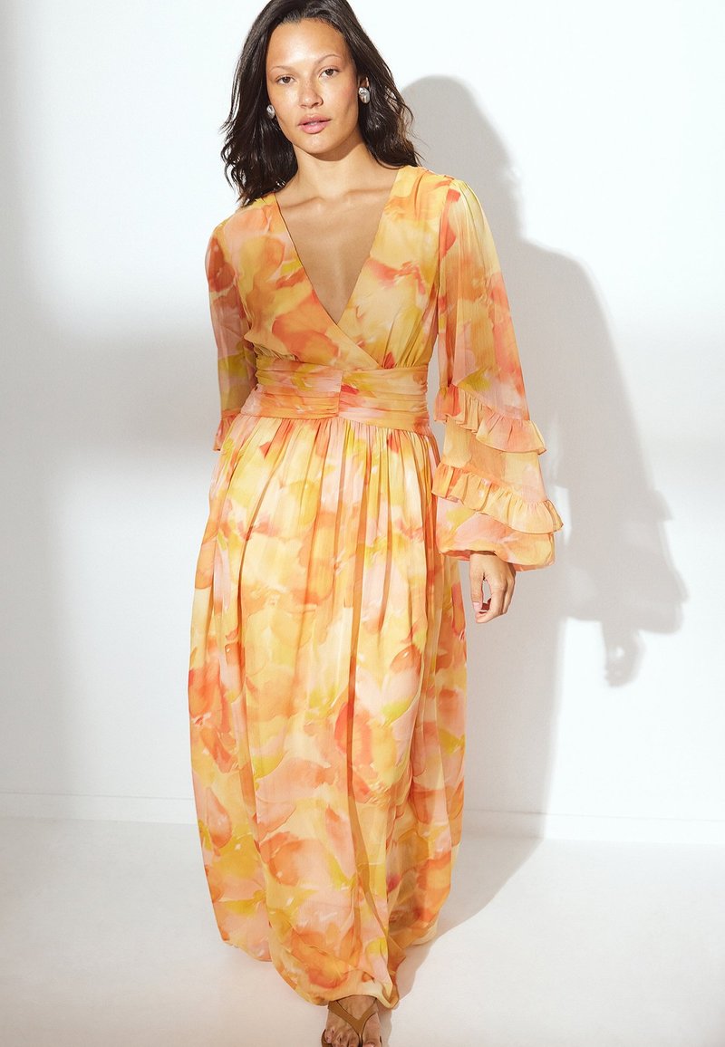 Frau in einem fließenden, langärmeligen Kleid mit tiefem V-Ausschnitt, das orangefarbene, gelbe und rosa Aquarellblumenmuster sowie Rüschen an den Ärmelabschlüssen aufweist.