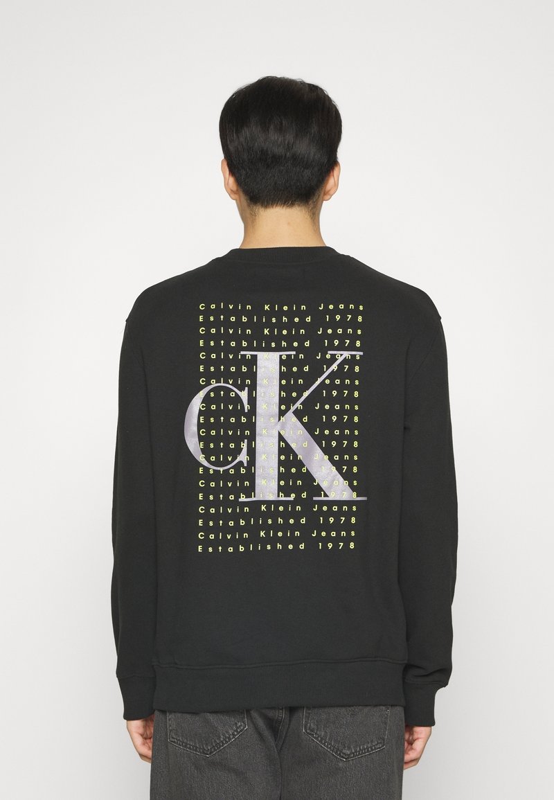 Homme portant un sweat noir avec le texte jaune répété "Calvin Klein Jeans Established 1978" et un grand logo argenté "CK" au dos.