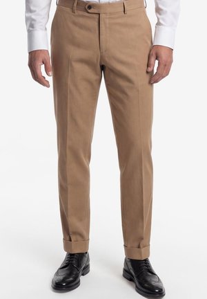 Mann trägt beige maßgeschneiderte Hose mit Umschlägen, schwarze Lederschuhe und ein weißes Hemd, steht vor weißem Hintergrund.