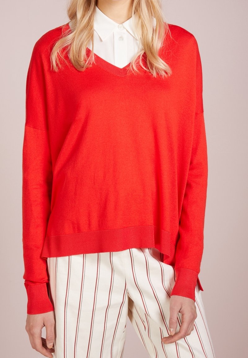 Femme portant un pull rouge vif à col en V sur une chemise blanche à col, associé à un pantalon blanc avec de fines rayures rouges verticales.