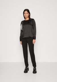Haut noir en satin à manches longues avec un accent floral, associé à un pantalon noir ajusté. Modèle portant des chaussures noires chunky, posant sur un fond uni.