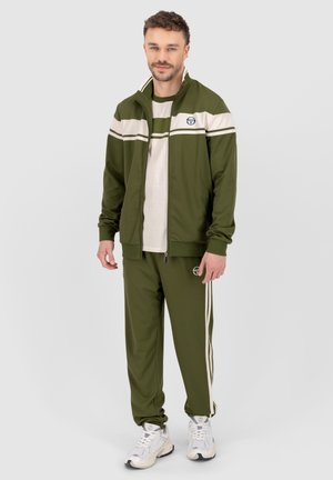 Olive green tracksuit with white stripes, zip-up jacket, logo on left chest, and matching pants with side stripes. White sneakers complete the look.