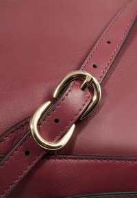 Bolso de mano de cuero marrón oscuro con una textura suave, que presenta un adorno metálico en tono dorado en la correa, resaltando el detalle de las costuras.