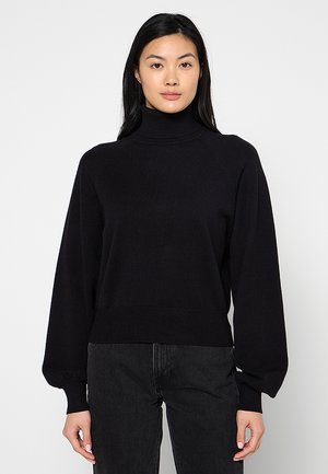 Kvinde med langt sort hår iført en sort rullekravetrøje og sorte bukser, stående mod en ensfarvet lys baggrund.