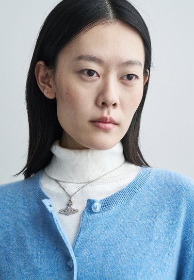 Cardigã azul claro feito de material suave, com gola redonda e botões, sobre uma camisola de gola alta branca, adornado com um colar decorativo.