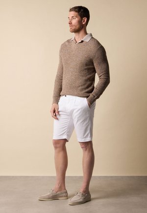 Mann trägt beigen Strickpullover über einem Hemd mit Kragen, weiße Shorts und beige Loafers, steht mit einer Hand in der Tasche vor einem schlichten Hintergrund.