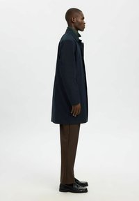 Marineblauer Mantel mit geradem Schnitt und Knopfverschluss, kombiniert mit einem grünen Rollkragenpullover, braunen Hosen und glänzenden schwarzen Schuhen, im Profil gestylt.