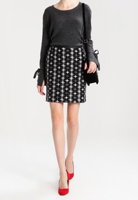 Maglione grigio scuro con maniche lunghe e dettagli a fiocco, abbinato a una gonna aderente con motivo bianco e nero e tacchi alti rossi.