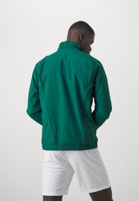 Chaqueta verde ligera con cuello alzado, cinturilla elástica y textura suave, que presenta un diseño simple y sin logotipos ni detalles visibles.
