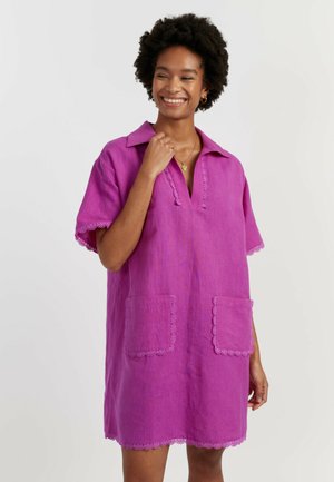 Donna sorridente con capelli corti e ricci che indossa un vestito viola ampio a maniche corte con bordatura in pizzo e tasche frontali su uno sfondo semplice.