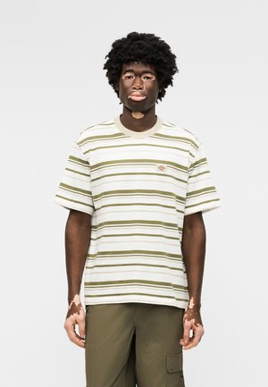 Νεαρός άνδρας με λεύκη που φοράει λευκό και ελαιώδες ριγέ μπλουζάκι και ελαιώδες παντελόνι cargo, στέκεται μπροστά σε απλό φωτεινό φόντο.