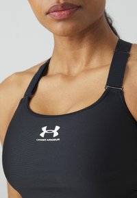 Crni sportski grudnjak napravljen od elastičnog materijala s podesivim trakama. Ima bijeli Under Armour logo sprijeda i gladak teksturu.