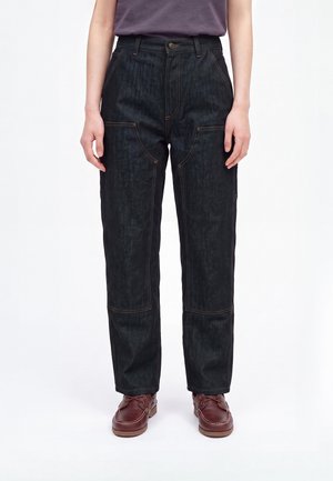 Person iført mørke denim bukser med lige ben og synlige syninger samt brune læder snørestøvler, stående mod en ensfarvet hvid baggrund.