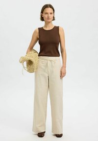 Brun ärmlös topp i kombination med ljusbeige randiga vida byxor. Modellen håller en vävd tan-väska. Strukturerad, avslappnad look.